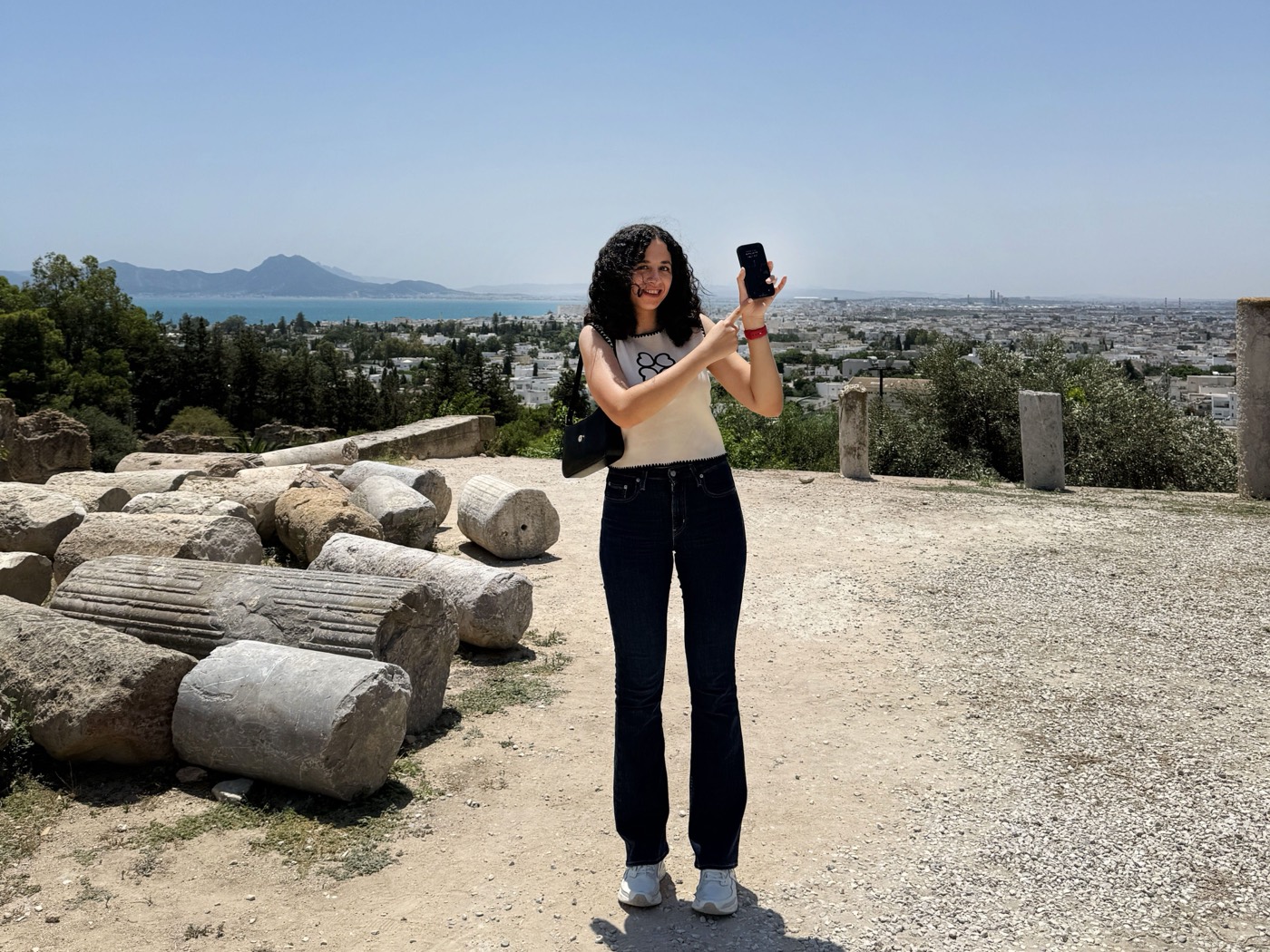 A person stands among ancient ruins in Tunisia, holding a smartphone and pointing towards the city landscape in the background.